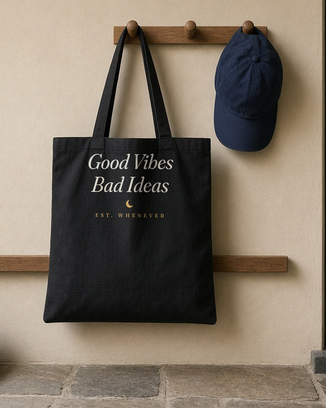 The House Tote in Black, black tote hanging on a peg by the front door beside a navy cap, shoes below