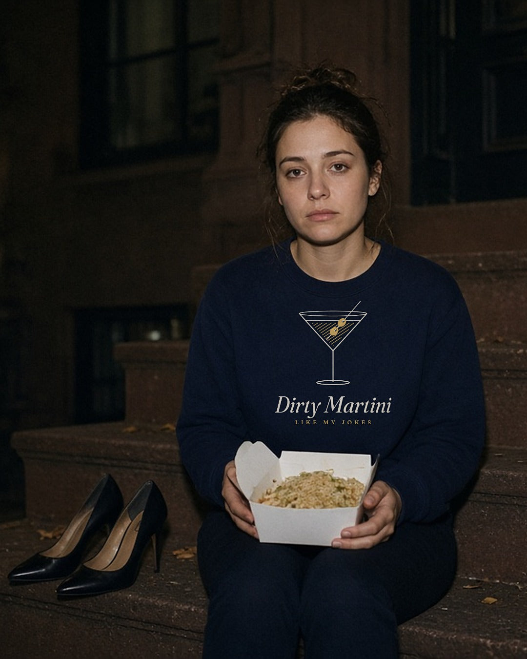 The Dirty Martini Crewneck in Navy, woman (late 20s) on brownstone steps at night with a takeout box, heels beside her, direct flash