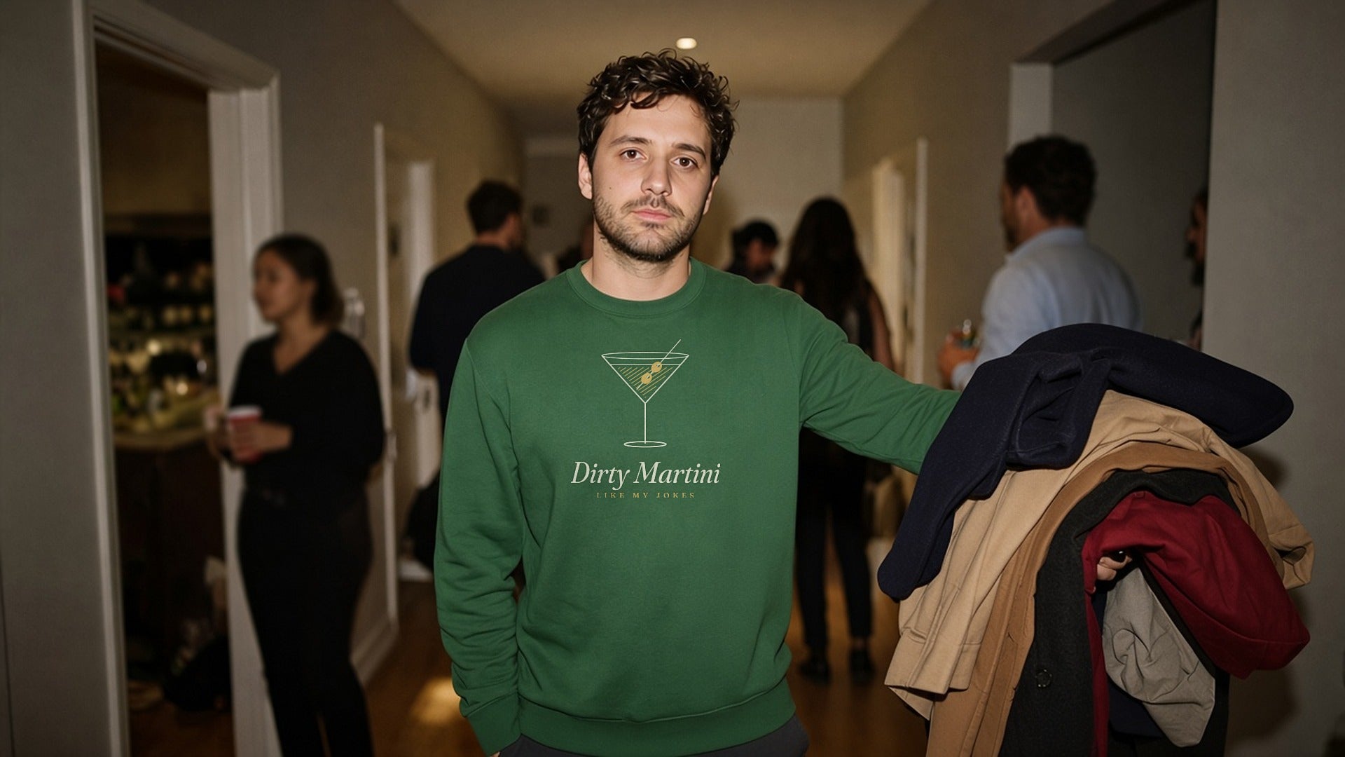 The Dirty Martini Crewneck in Forest Green, man in the Forest Green crewneck holds an armful of coats at a house party, guests behind, flash