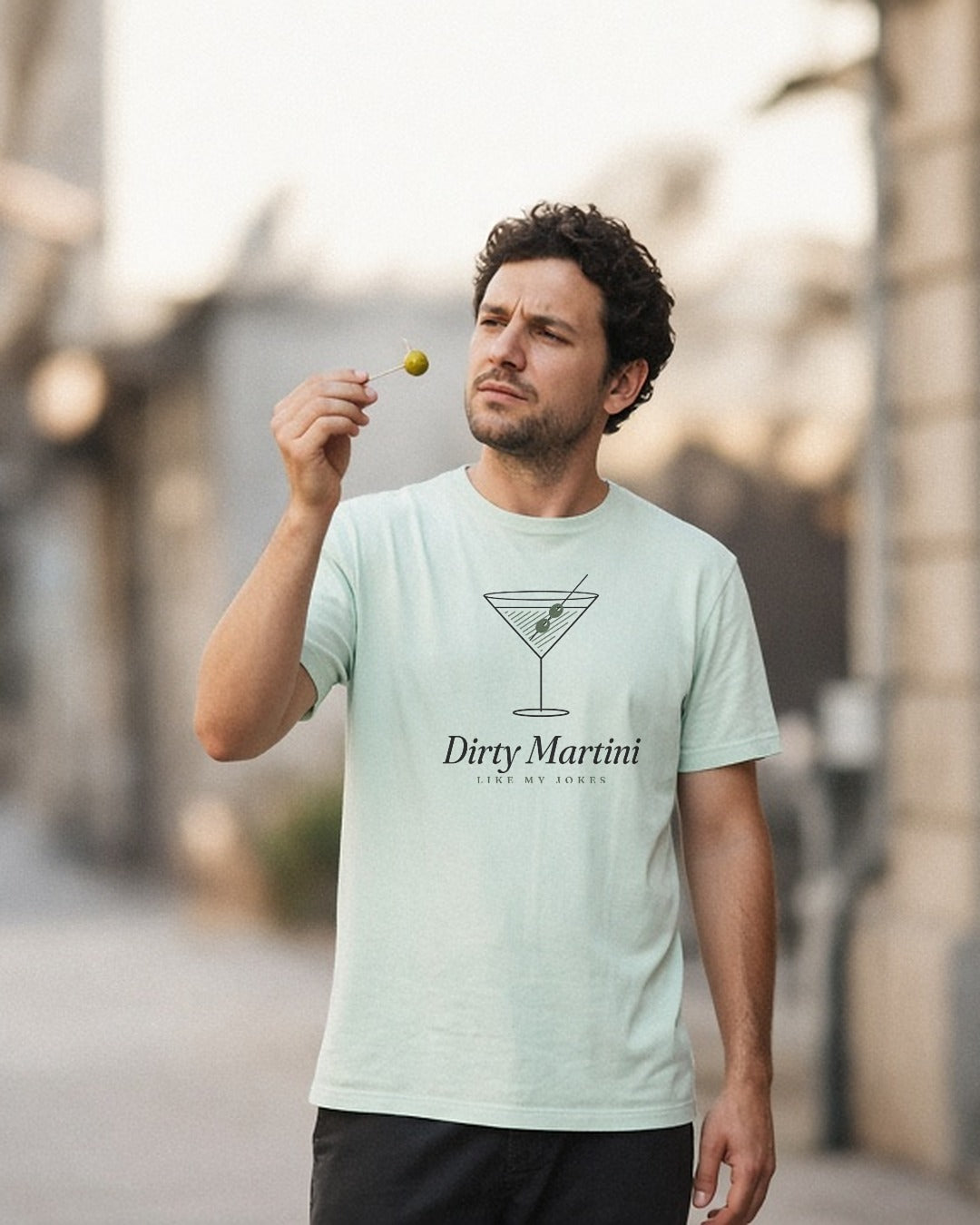 The Dirty Martini Tee in Chalky Mint, man holds a single olive up to the light like a jeweler, city street, open shade
