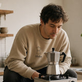 The Moka Crewneck in Sand, worn by a man waiting on a moka pot over a camp burner