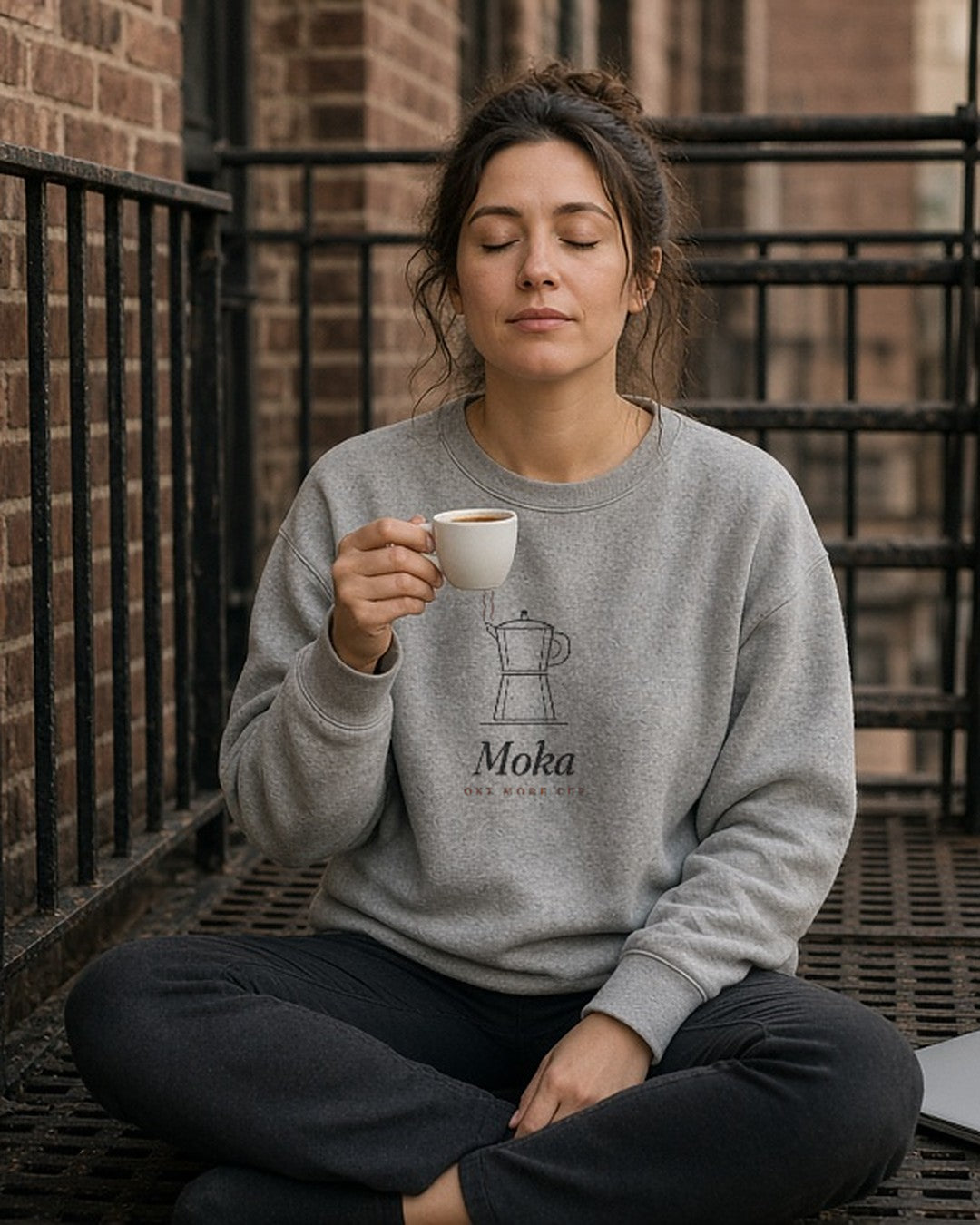 The Moka Crewneck in Ash, worn by a woman sitting cross-legged on a fire escape with an espresso