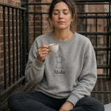 The Moka Crewneck in Ash, worn by a woman sitting cross-legged on a fire escape with an espresso