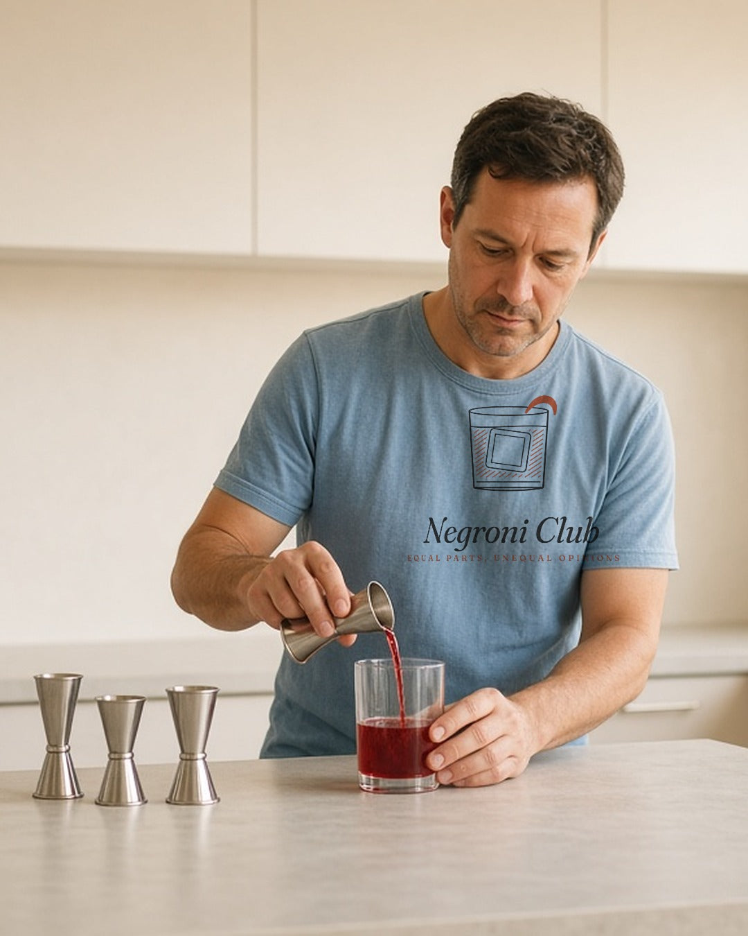 The Negroni Club Tee in Chambray, man (40s) in the Chambray tee carefully pours from three small jiggers on a kitchen counter, window light