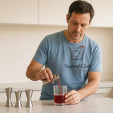 The Negroni Club Tee in Chambray, man (40s) in the Chambray tee carefully pours from three small jiggers on a kitchen counter, window light