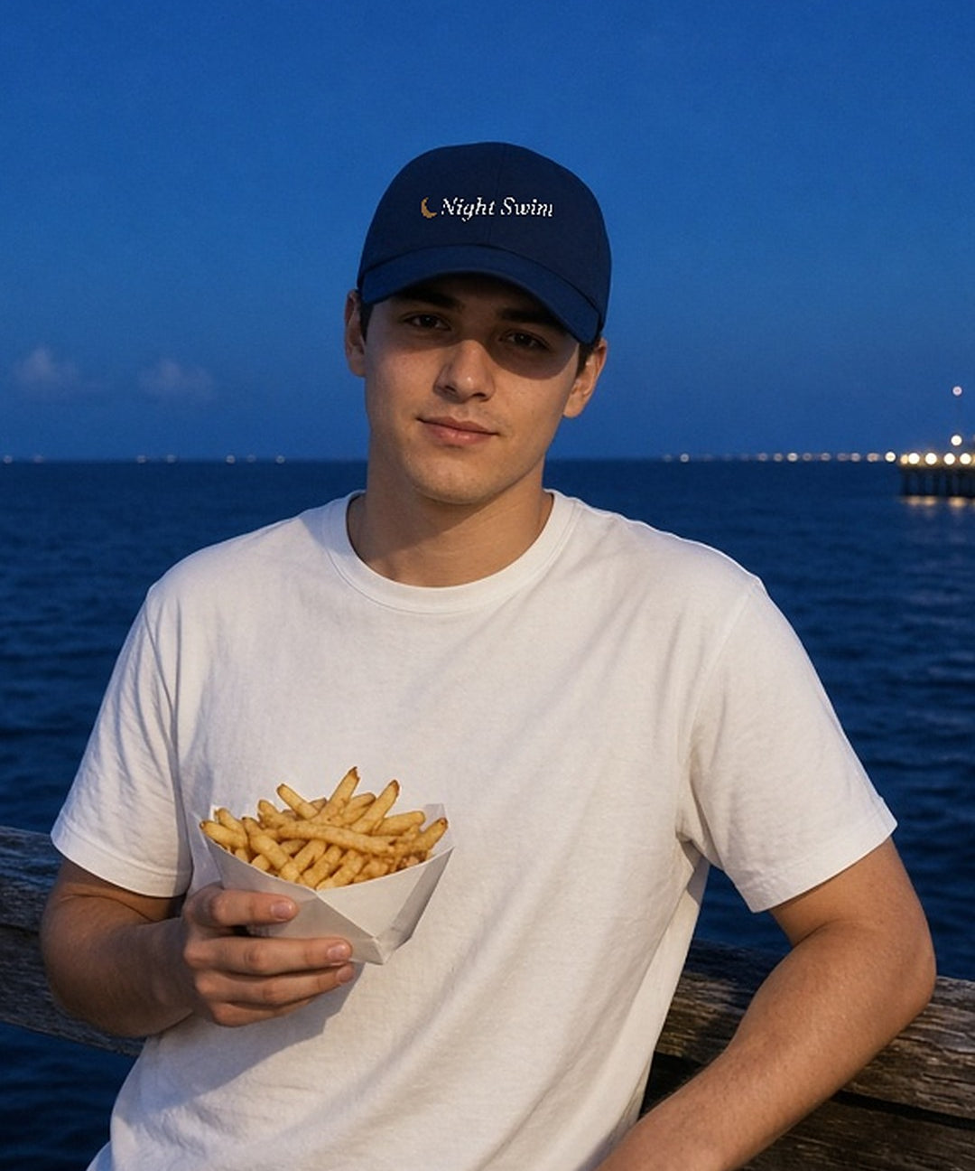 The Night Swim Dad Hat in Navy, worn at a boardwalk railing at blue hour with a boat of fries