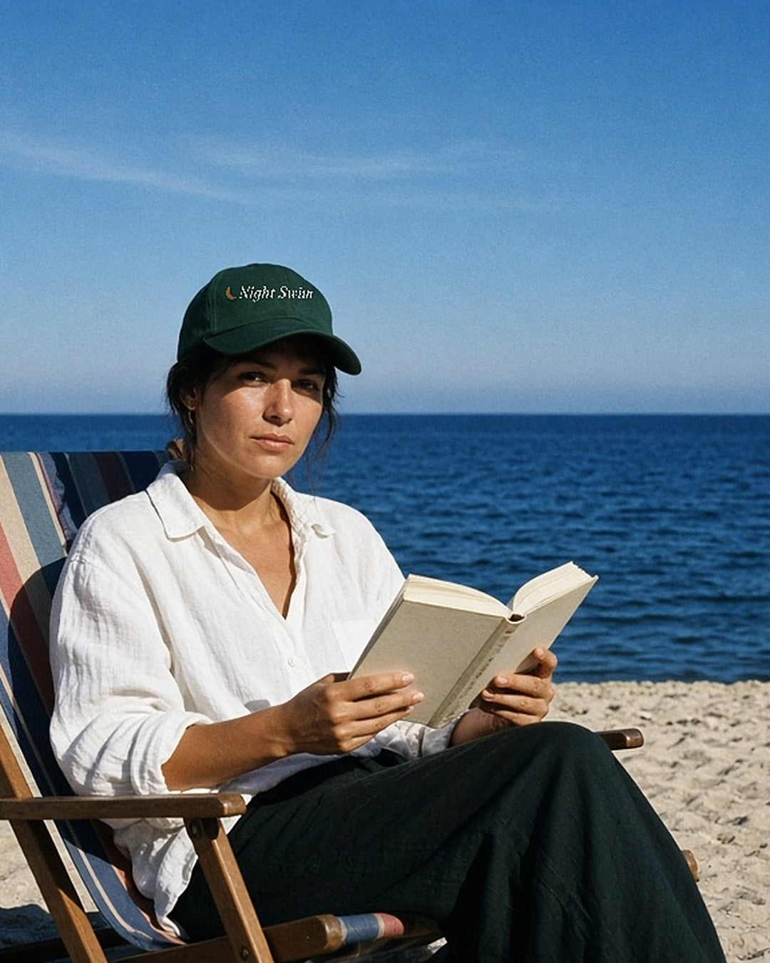The Night Swim Dad Hat in Spruce, worn by a woman reading in a striped deck chair
