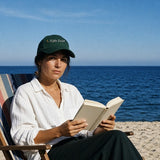 The Night Swim Dad Hat in Spruce, worn by a woman reading in a striped deck chair
