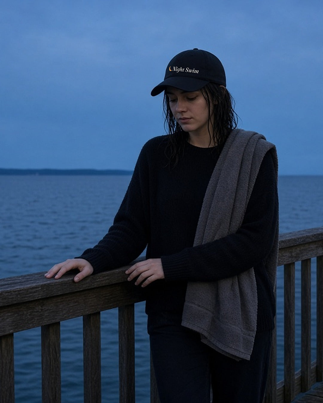 The Night Swim Dad Hat in Black, young woman with wet hair and a towel over her shoulder leaning on a pier rail at dusk