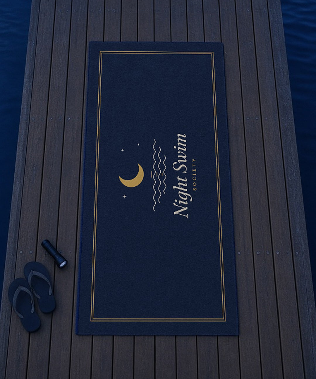 The Night Swim Beach Towel laid out on a dock with flip-flops and a flashlight, seen from above