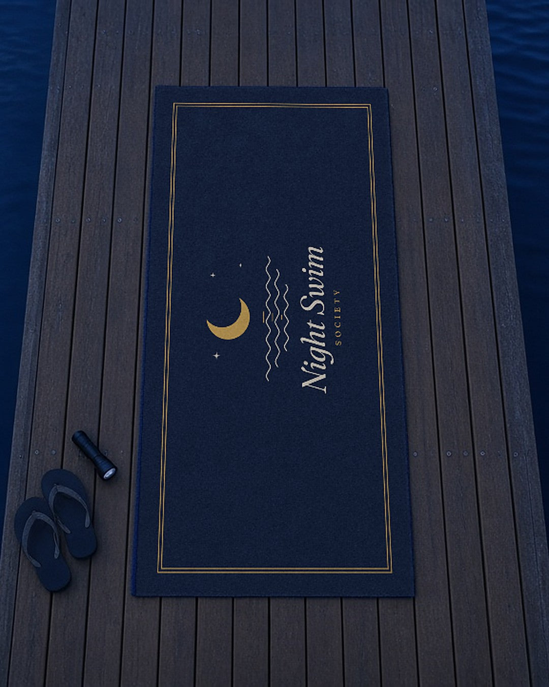The Night Swim Beach Towel laid out on a dock with flip-flops and a flashlight, seen from above