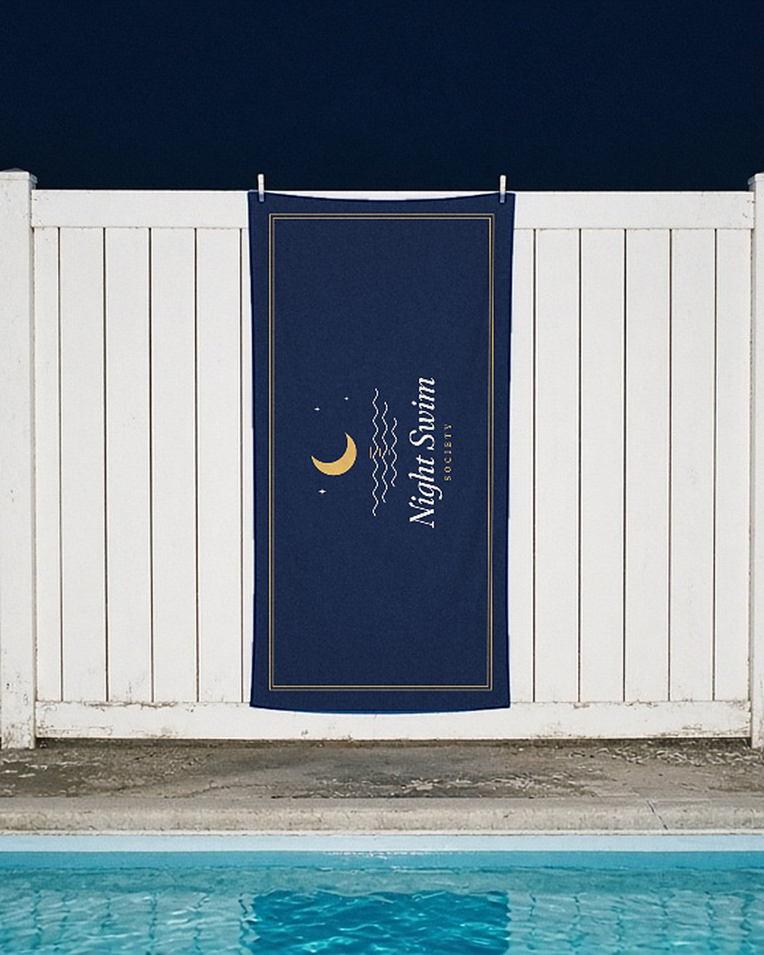 The Night Swim Beach Towel clipped to a white fence beside a motel pool at night