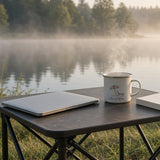 The Out of Office Camp Mug on a camp table by a misty lake next to a closed laptop