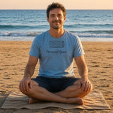 The Personal Space Tee in Chambray, man in the Chambray tee alone, cross-legged on a tiny towel on a wide sandy beach, sea behind