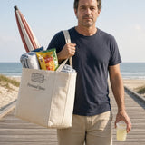 The Personal Space Tote in Oyster, man on a beach boardwalk with the tote on his shoulder, a beach umbrella, towel and snacks packed, drink in hand
