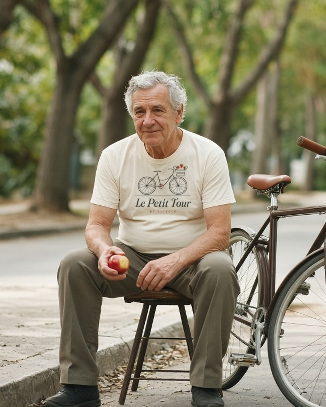 Le Petit Tour Tee in Ivory, a man (60s) in the Ivory tee calmly sitting on a curb next to his bike with a flat, eating an apple
