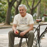 Le Petit Tour Tee in Ivory, a man (60s) in the Ivory tee calmly sitting on a curb next to his bike with a flat, eating an apple