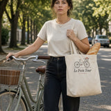 Le Petit Tour Tote in Oyster, woman walking her bike down a tree-lined street, tote on her shoulder with a baguette