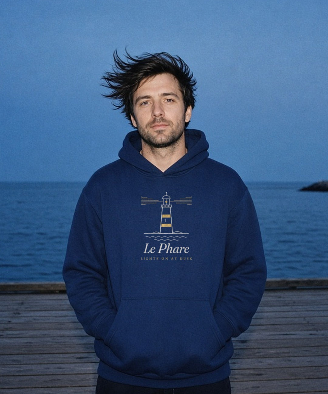 Le Phare Hoodie in Navy Blazer, a man (30s), hood down, hands in the pocket, hair blowing sideways on a pier at blue hour