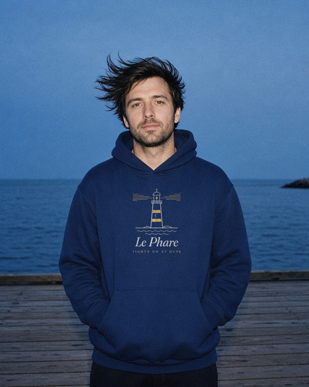 Le Phare Hoodie in Navy Blazer, a man (30s), hood down, hands in the pocket, hair blowing sideways on a pier at blue hour