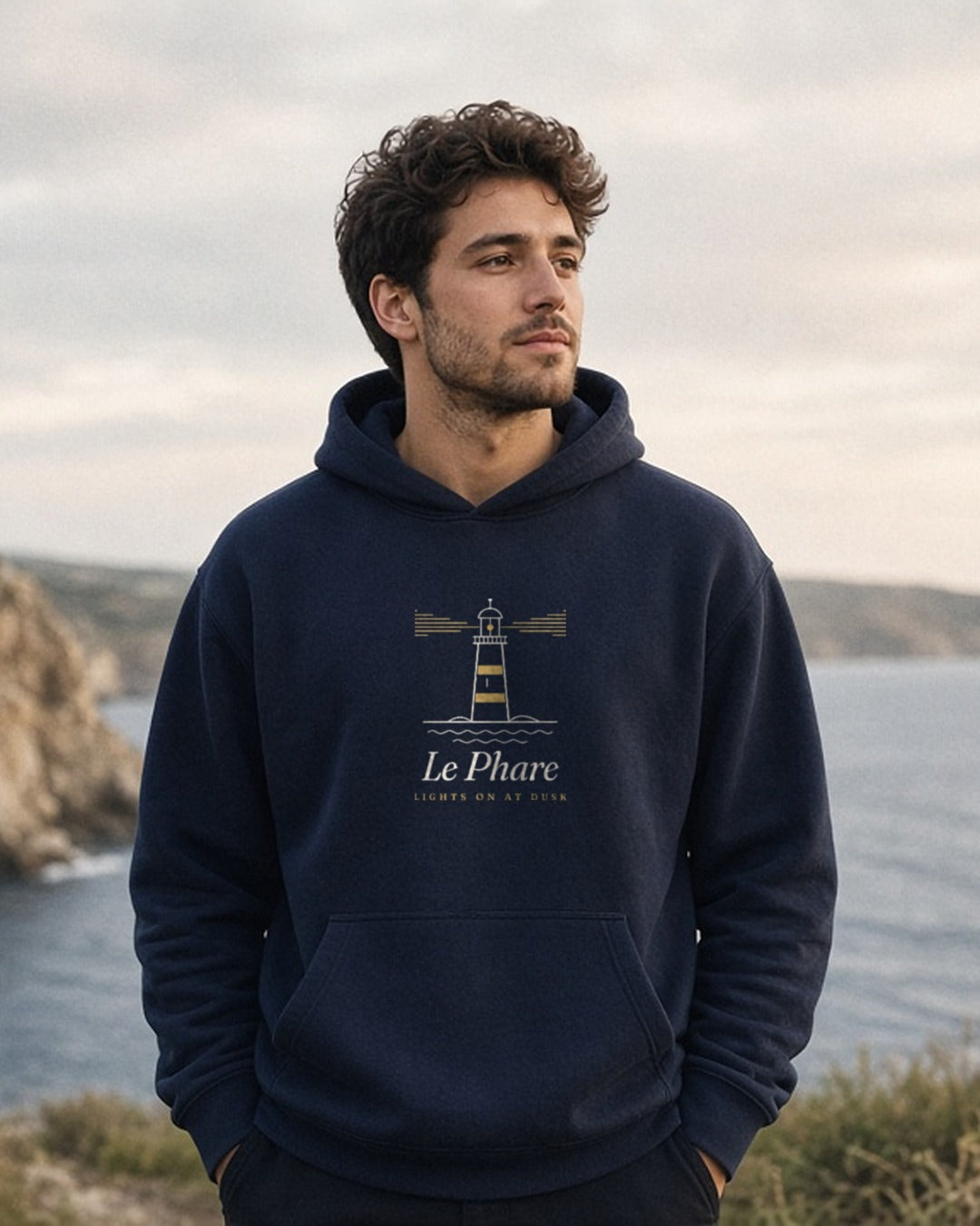 Le Phare Hoodie in Navy Blazer, man on a coastal clifftop path, overcast evening, hands in the hoodie pocket