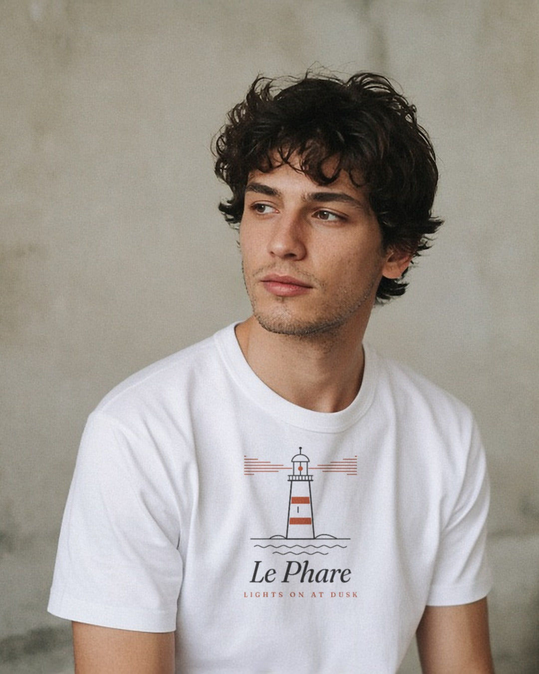 Le Phare Tee in White, close portrait of a young man in a white tee against a pale concrete wall, looking off to the side