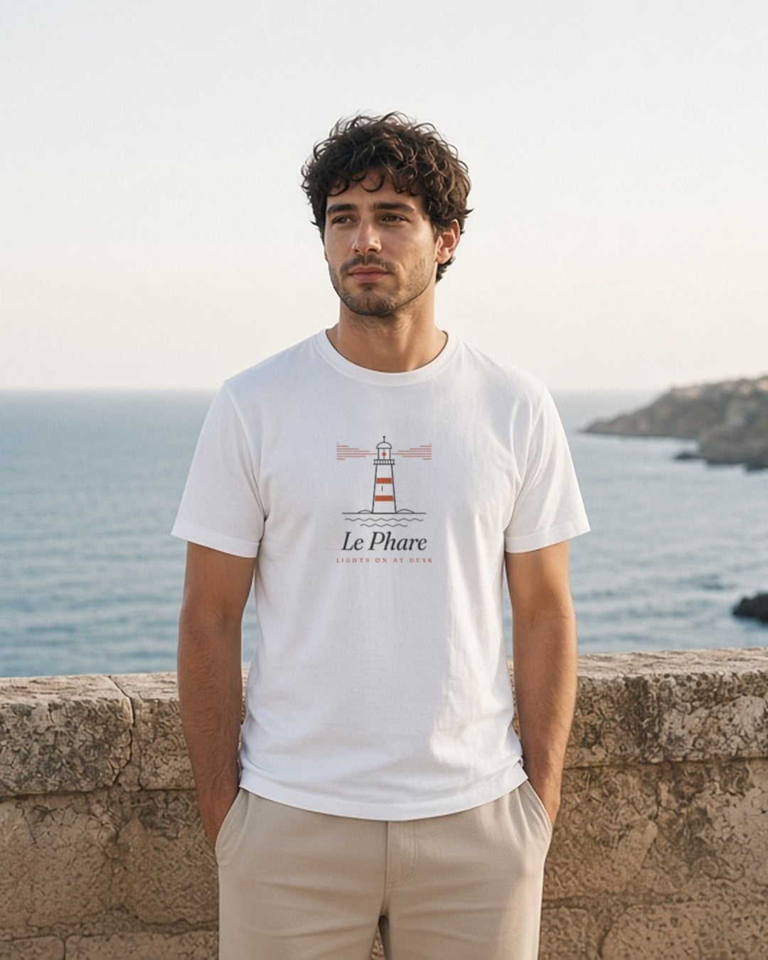 Le Phare Tee in White, man standing at a stone seawall above a rocky coast, hands in pockets