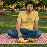 The Picnic Club Tee in Butter, man in the Butter tee alone, cross-legged on a red gingham blanket in a park with a cheese board in front