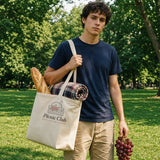 The Picnic Club Tote in Oyster, young man in a park, tote on his shoulder with a baguette and a picnic blanket, bunch of grapes in hand