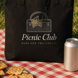 The Picnic Club Tote in Black, the Black tote on a picnic blanket, cheese, crackers and soda cans around it