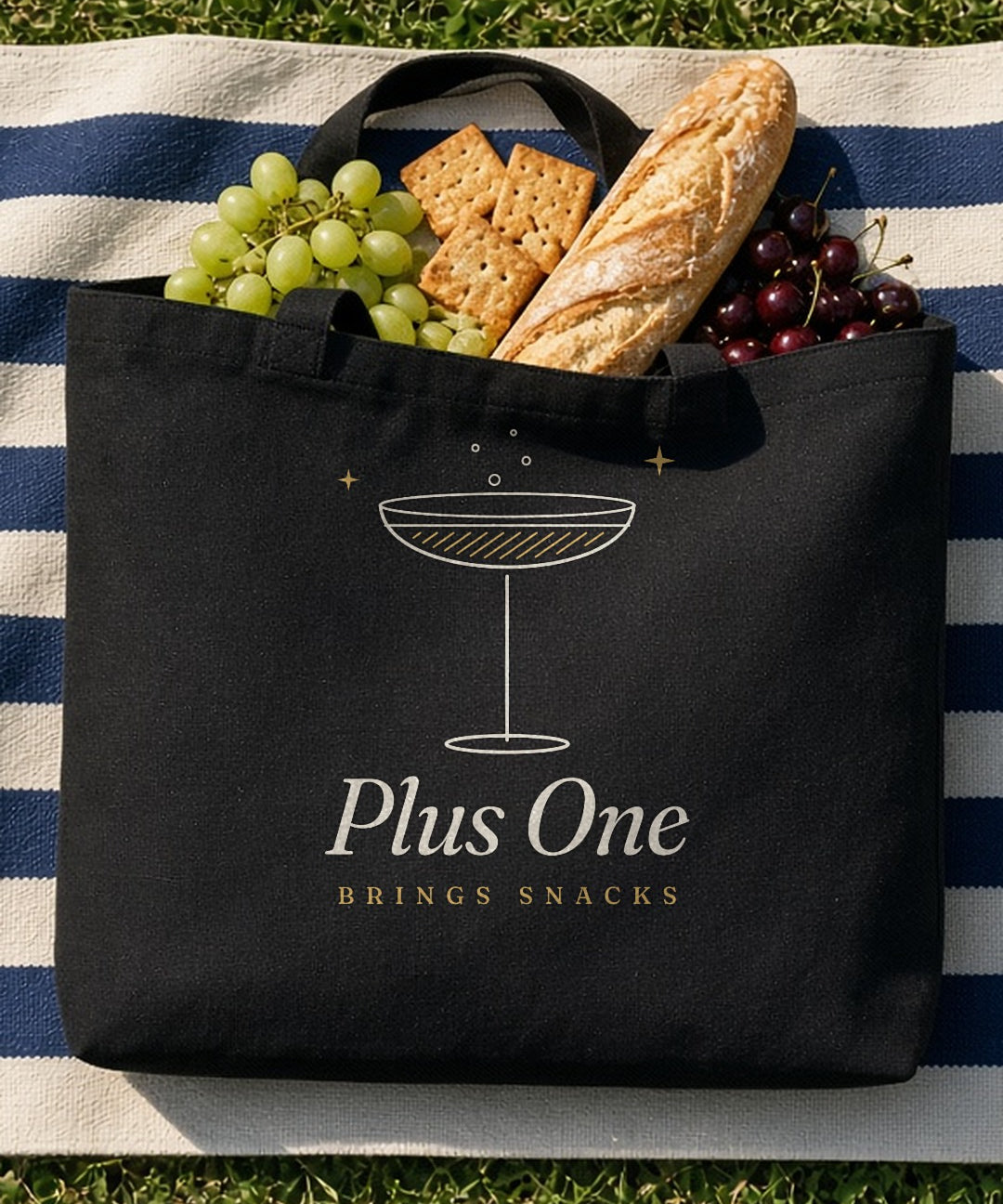 The Plus One Tote in Black, black tote on a navy-striped blanket on grass, baguette, crackers and grapes spilling out, overhead