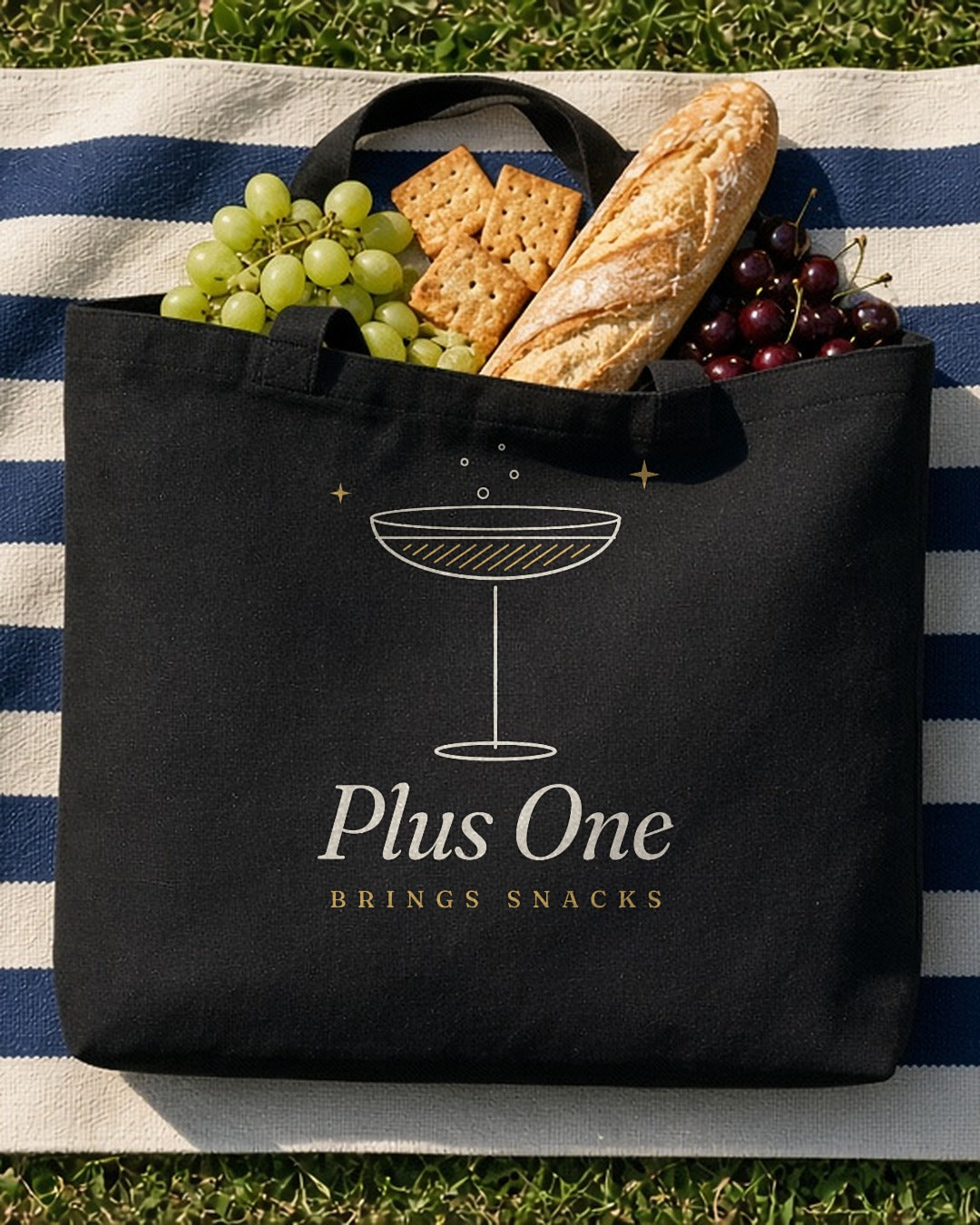The Plus One Tote in Black, black tote on a navy-striped blanket on grass, baguette, crackers and grapes spilling out, overhead