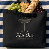 The Plus One Tote in Black, black tote on a navy-striped blanket on grass, baguette, crackers and grapes spilling out, overhead