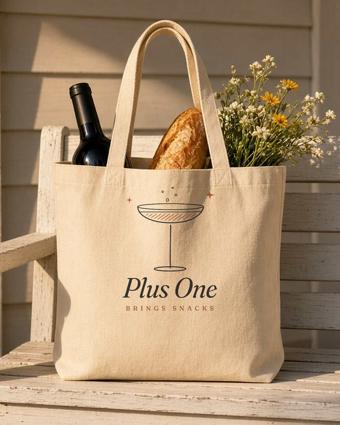 The Plus One Tote in Oyster, tote on a porch packed with a bottle of wine, a baguette and wildflowers
