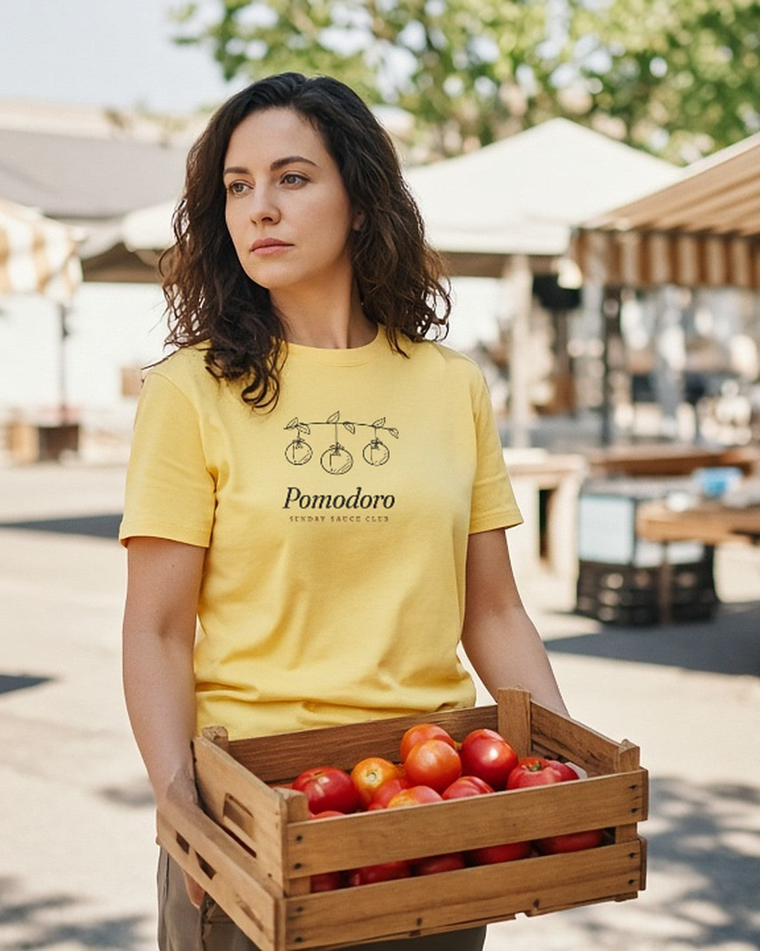 The Pomodoro Tee in Butter, worn by a woman carrying a crate of tomatoes at a farmers market