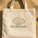 The Sea You Later Tote in Oyster, the tote on the sand with shells and a striped towel