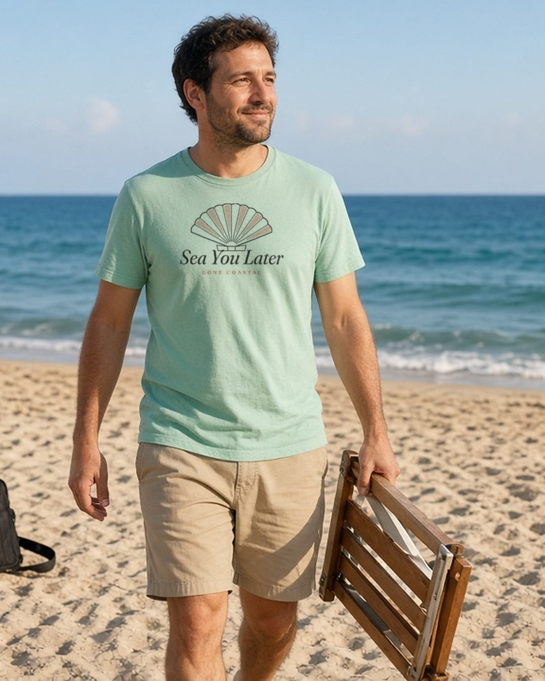 The Sea You Later Tee in Chalky Mint, a man (30s) walking along the beach carrying a folded wooden beach chair