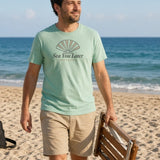 The Sea You Later Tee in Chalky Mint, a man (30s) walking along the beach carrying a folded wooden beach chair