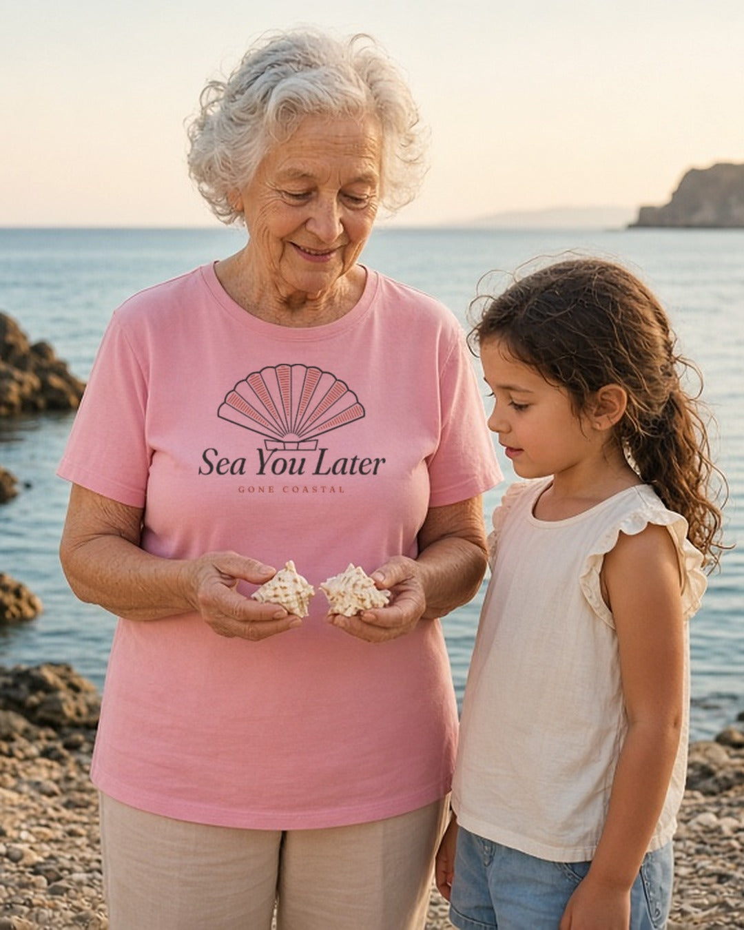 The Sea You Later Tee in Blossom, grandmother showing seashells to her granddaughter on a rocky shore at golden hour