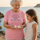 The Sea You Later Tee in Blossom, grandmother showing seashells to her granddaughter on a rocky shore at golden hour