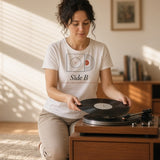 The Side B Tee in White, woman cross-legged on a rug flips a record on a turntable, sun streaks through the blinds