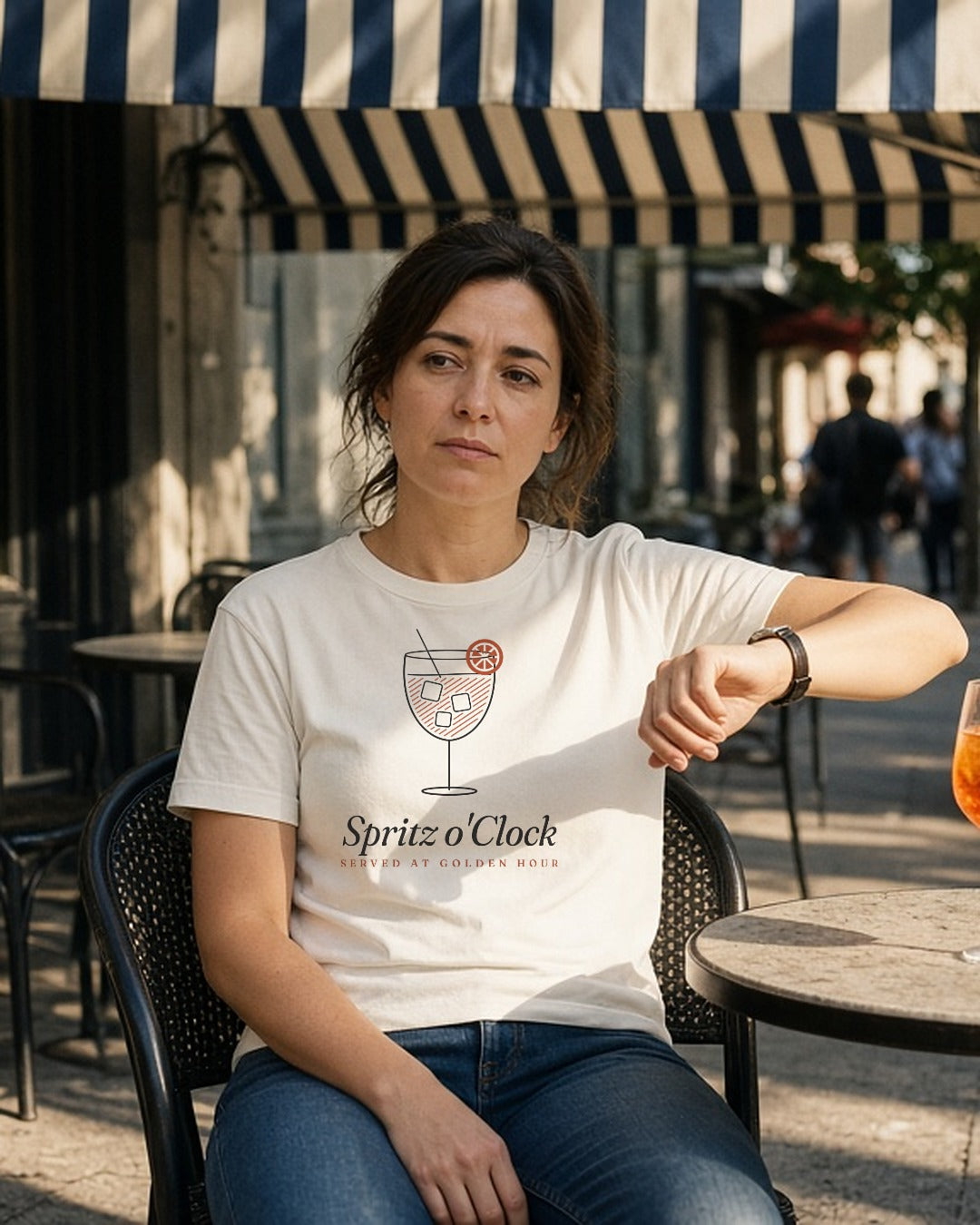 The Spritz o'Clock Tee in Ivory, woman (30s) sits back in a café chair on a sunny sidewalk under a navy/white striped awning, raising her watch wrist beside her chest to check the time; orange spritz with an orange slice untouched on the marble table; hard late sun