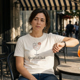 The Spritz o'Clock Tee in Ivory, woman (30s) sits back in a café chair on a sunny sidewalk under a navy/white striped awning, raising her watch wrist beside her chest to check the time; orange spritz with an orange slice untouched on the marble table; hard late sun