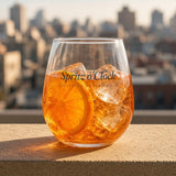 The Spritz o'Clock Stemless Glass in Clear 15 oz, glass with orange spritz and orange slice on a sun-warmed ledge, city rooftops behind, hard sun