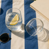 The Spritz o'Clock Stemless Glass in Clear 15 oz, two glasses on a blue-striped blanket, one upright with sparkling water and lemon, one knocked over (empty), overhead