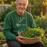 The Thyme Flies Crewneck in Forest Green, a man (60s) in Forest Green on a garden stool with a basket of cut herbs, golden low sun
