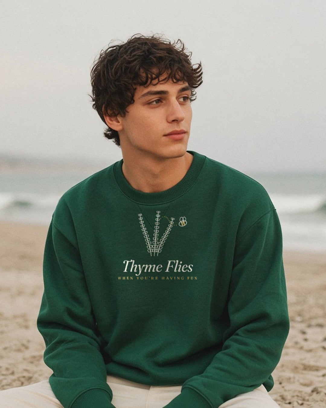 The Thyme Flies Crewneck in Forest Green, young man in a Forest Green crew sitting alone on a beach on an overcast day, looking off to the side