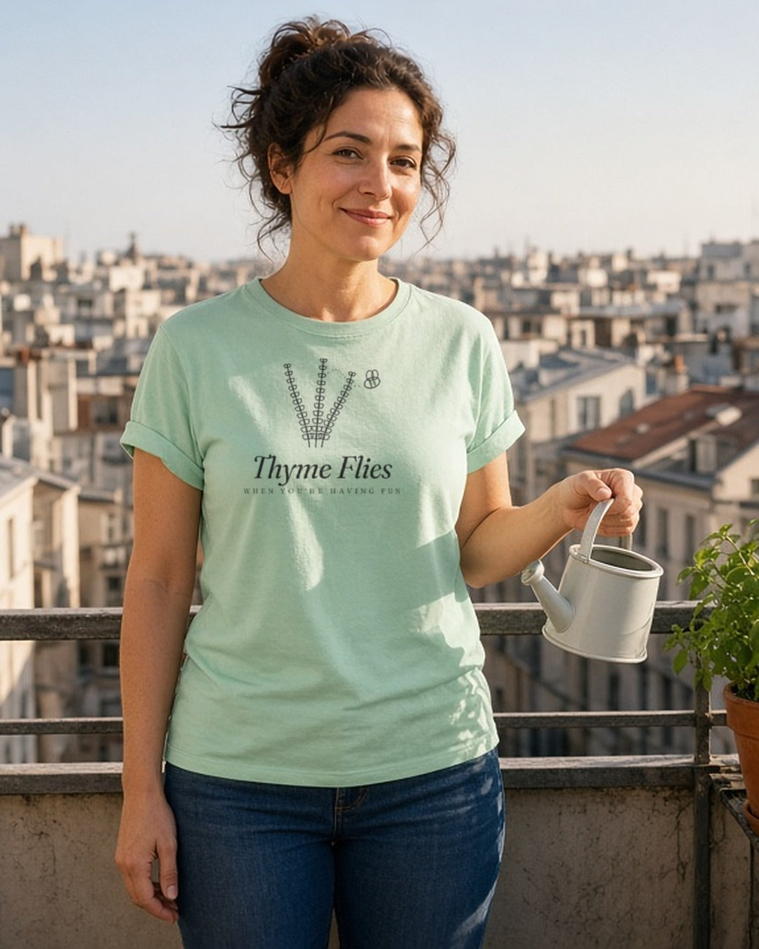 The Thyme Flies Tee in Chalky Mint, a woman (30s) in the Chalky Mint tee on a tiny apartment balcony proudly watering a single pot of herbs