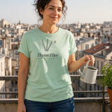 The Thyme Flies Tee in Chalky Mint, a woman (30s) in the Chalky Mint tee on a tiny apartment balcony proudly watering a single pot of herbs