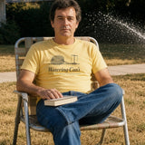 The Watering Can't Tee in Butter, a man (40s) in a lawn chair reading a paperback while a sprinkler waters the sidewalk instead of the yellow lawn